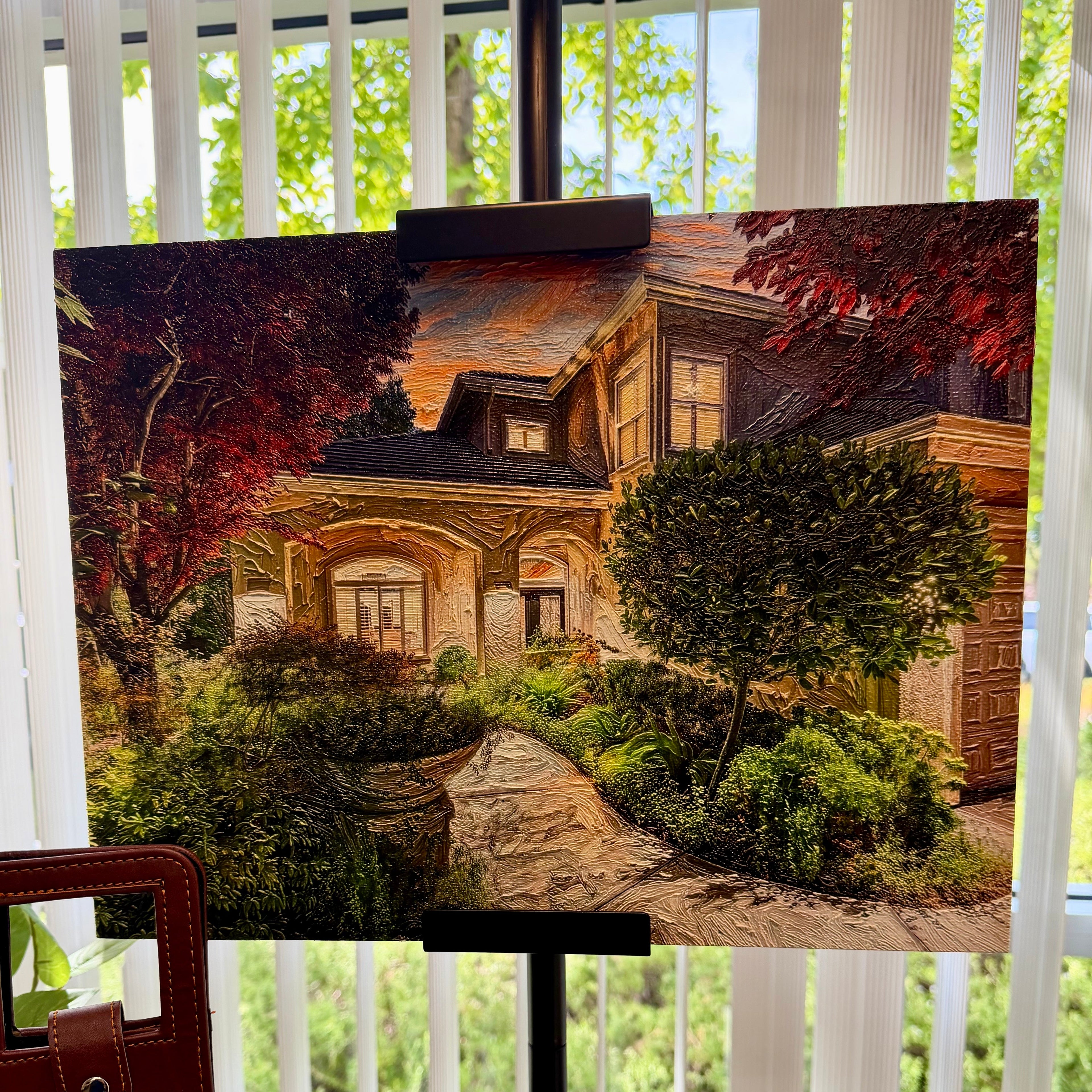 A canvas painting of a house with a stone path and greenery, displayed on a black easel in front of a window with white vertical blinds.