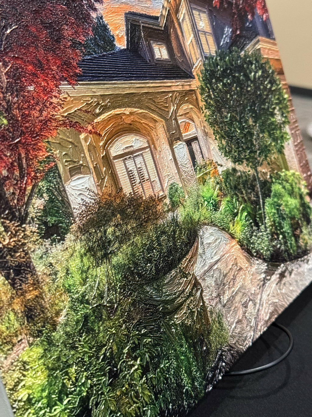 A close-up of a canvas print or panel featuring a detailed painting of an old stone house with a garden path, surrounded by greenery and red autumn foliage.